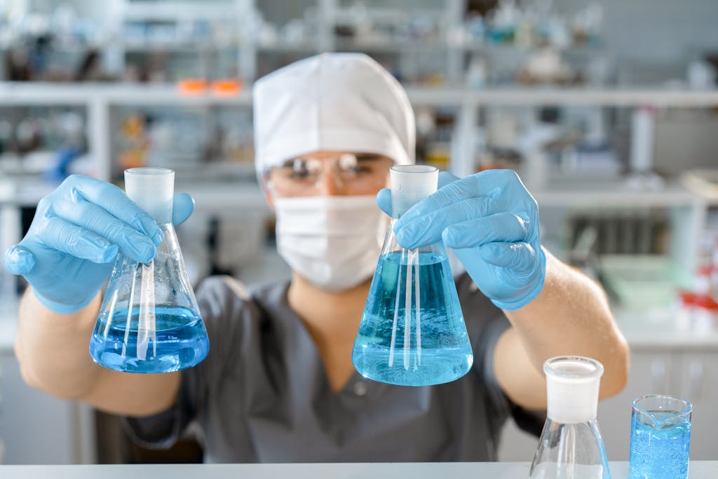 A scientist in protective gear handling blue liquids in laboratory flasks.